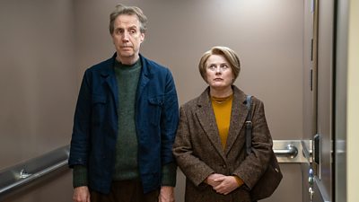 Scene from Onwards and Sideways. Tony Evans (Rhys Ifans) and Liz Evans (Monica Dolan) stand in an elevator.