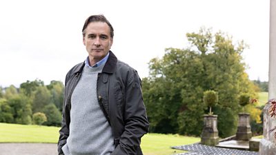 A man in a blue shirt and grey jumper stands outdoors, wearing a dark coat as he looks to camera. A large lawn is visible behind him.