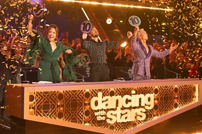 Dancing with the Stars US judges (Carrie Ann Inaba, Derek Hough and Bruno Tonioli) on their feet holding padels with 10s on.
