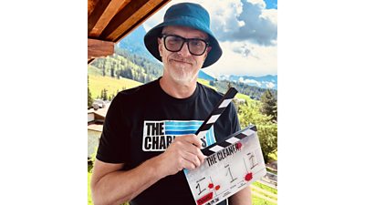 Greg Davies holds a clapperboard for a new episode of The Cleaner. He stands outdoors in the Italian mountains.