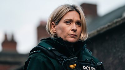Siân Brooke in character as Grace Ellis. Dressed in police-branded protective gear, she stands outside and looks at something offscreen.