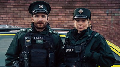 Stevie Neil in character as Martin McCann and Siân Brooke in character as Grace Ellis. Both are dressed in police-branded protective gear and stand in front of a police car.