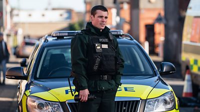Nathan Braniff in character as Constable Tommy Foster. Dressed in police-branded protective gear, he stands in front of a police car on a residential street.
