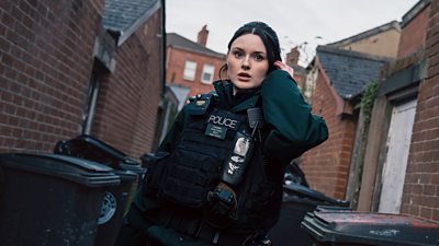 Katherine Devlin in character as Constable Annie Conlon. Dressed in police-branded protective gear, she stands in an alleyway in between houses. Looking at someone offscreen in concern, she reaches to her earpiece.