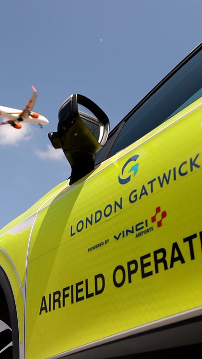 A yellow airfield operation car with a plane in the sky.
