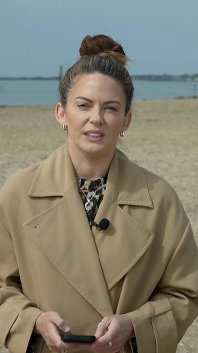 Female reporter with brown tied-up hair and brown coat stands on beach