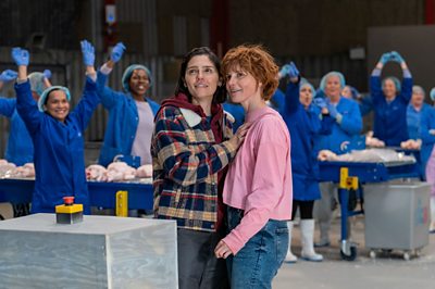 A scene from Chuck Chuck Baby. Two people react in surprise in the centre of the image. In the background, lots of factory works in blue overalls and blue hairnets celebrate with their arms raised.