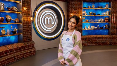 A woman in a green and pink striped jumper and a white apron standing in the masterchef studio