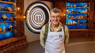 A man in a green shirt and a white apron stood in the masterchef studio smiling towards camera
