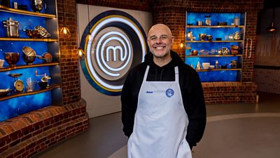 A man wearing a black hoodie and a white apron stood in the masterchef studio smiling towards the camera