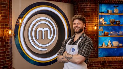 A man in a plaid shirt and a white apron stood in the Masterchef studio smiling towards camera
