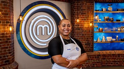 A woman in a black t-shirt and white apron stood in the masterchef studio smiling towards camera