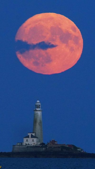 An orange-red full Moon, slightly obscured by cloud, above a lighthouse