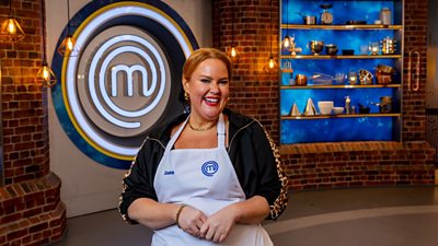 A woman wearing a black jacker and a white apron is standing in the masterchef studio smiling towards the camera