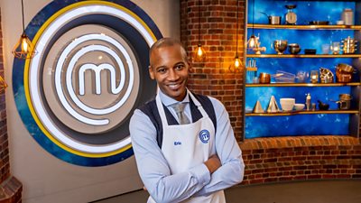 A man in a blue shirt, waistcoat and tie stands with his armed crossed, smiling towards camera in the masterchef studio
