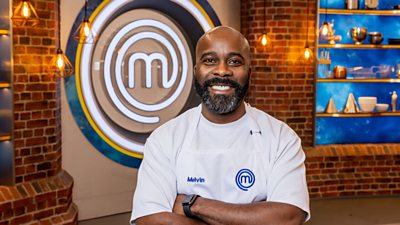 A man wearing a white t-shirt and white apron. He is standing with his arms crossed, smiling, in the masterchef studio