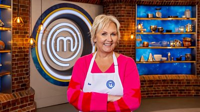 A woman in a bright pink shirt and white apron standing in the masterchef studio smiling