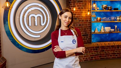 A woman in a red shirt and white apron standing in the masterchef studio