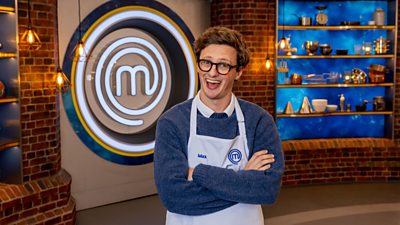 A man in a blue jumper and glasses crossing his arms and laughing in the masterchef studio
