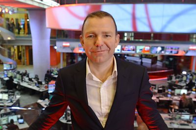 A man looks at the camera against a newsroom in background