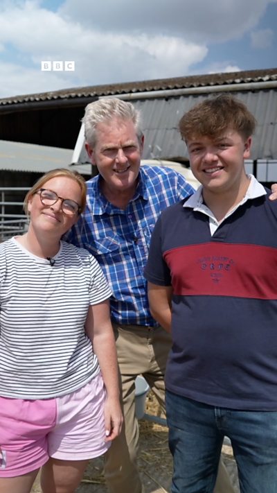 Two young farmers, one woman, one man, standing in front of a barn with a male reporter in the middle of them.