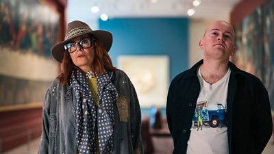 A woman and a man stood in an art gallery looking past the camera