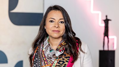Cordelia de Freitas in a white jacket and multi-coloured scarf looking into camera while stood in a gallery