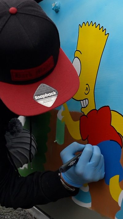 A person wearing a black and red cap uses a marker to draw on a colourful mural