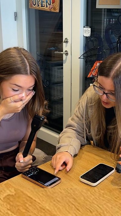 Two girls sitting at a table, one pointing at a phone screen and the other covers her mouth with her hand.