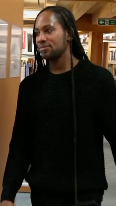 Jason Arday, dressed in black, walks through a library