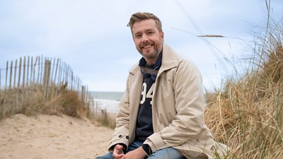 Iain Stirling smiling at the camera well sat against a grassy sand dune on a beach