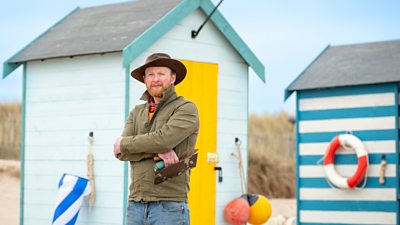 Jamie Wardley wearing a green jacket and a hat stood with his armed crossed stood in front of some brightly coloured blue beach huts