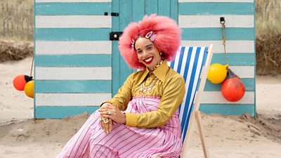 Sadie Clayton in a pink skirt and a yellow shirt sat on a striped deck chair on a beach in front of a striped beach hut