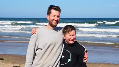 David and Zara stood on the beach with their arms around each others shoulder while stood on a beach in front of the sea