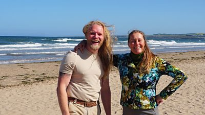 Alex Burman and Rémy Hoggard laughing and smiling towards camera while stood on a beach with the sea in the background