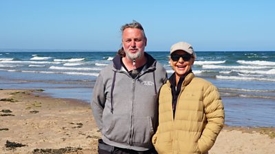 A man and a woman stood side by side smiling into camera while stood on a beach in Scotland
