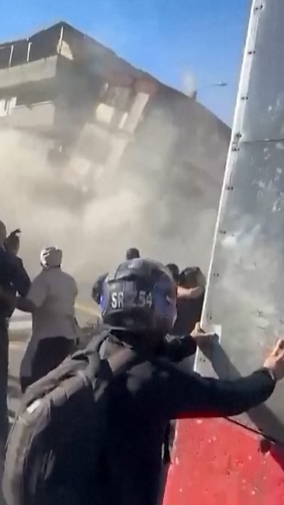 A building mid-collapse. A man holds on to a metal structure in the forefront.