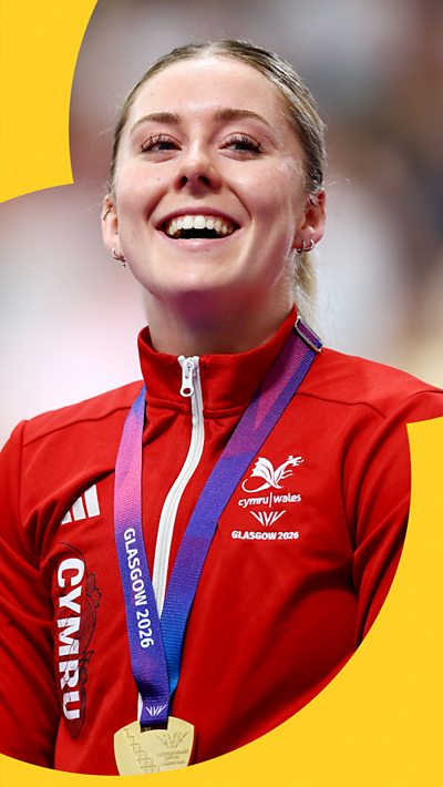 Welsh cyclist Emma Finucane smiling. She is wearing a red Wales tracksuit and has a gold medal around her neck.