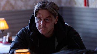 Scene from This City is Ours series 2. Banksey (Mike Noble) wears a large coat and sits at a table in a restaurant. He has an injury around his eye. 