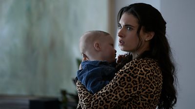Scene from This City is Ours series 2. Melissa (Darci Shaw) holds a baby and looks in shock at something offscreen. 