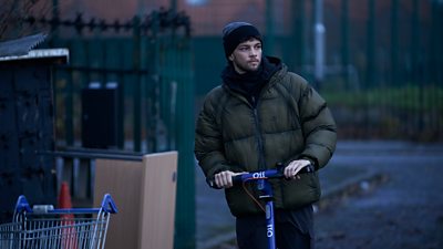 Scene from This City is Ours series 2. Freddie (Adam Abbou) using a scooter on a street. Furniture and abandoned items including traffic cones and a shopping trolley appear on the side of the road. 