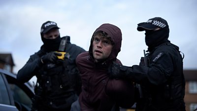 Scene from This City is Ours series 2. Bonehead (Bobby Schofield) is held by two masked police officers. They hold his arms behind his back as they walk outside beside a car. One of the officers holds onto a yellow taser with his free hand. 