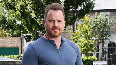 A man in a blue henley t-shirt with a short haircut and short beard stood in Albert Square looking into the camera 
