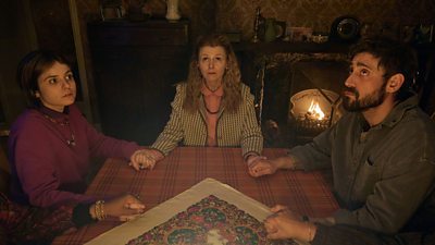 Three people sat at a wooden table in a dark room, holding hands and looking eerily upwards