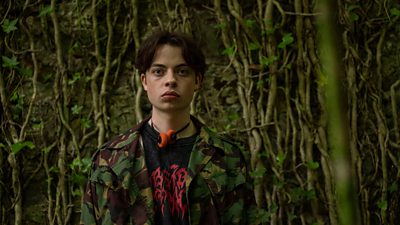A teenaged boy in a black t-shirt, camo jacket, and headphones. He's stood against a rock wall with tree roots dangling down
