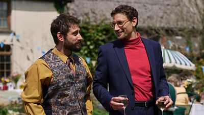 Two smartly dressed men in shirts and jackets with one holding two glasses of wine, they are stood in the garden of a country cottage