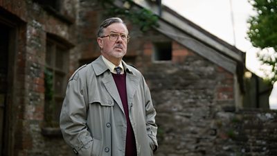 A man with grey hair and a stubbly grey beard and glasses stands outside an old brick house in a beige trench coat