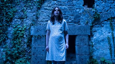 A woman in a white night dress stood outside a some ancient ruins with markings on a stone entrance