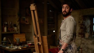 A man in beige t-shirt and white paint-covered apron stood next to an easel surrounded by paint