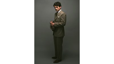 First look photo of Edward Bluemel as Hercule Poirot. Dressed in a suit and tie, he holds a bowler hat in both hands and looks to the camera. 